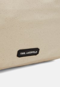 Beżowa torba z płótna o fakturowej powierzchni z czarną łatą z logo, na której widnieje "KARL LAGERFELD" w białym tekście. W kształcie sześciokąta.
