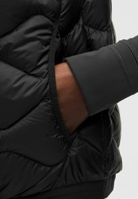 Musta toppatakki, jossa on tikattu kuvio ja sivutasku. Hihassa on sileä, istuva mansetti tummemmassa harmaan sävyssä.