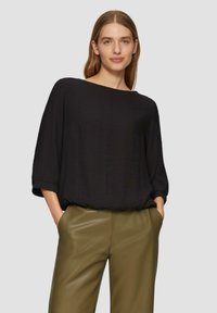 Haut noir ample à manches trois-quarts et décolleté bateau, associé à un pantalon taille haute vert olive. Texture de tissu lisse.