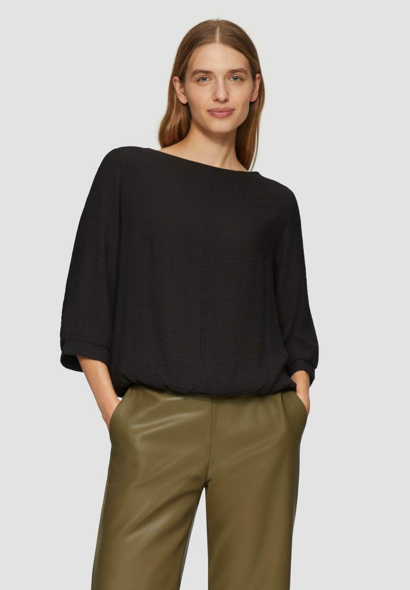 Haut noir ample à manches trois-quarts et décolleté bateau, associé à un pantalon taille haute vert olive. Texture de tissu lisse.