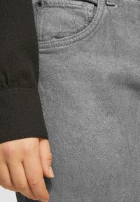 Grijze denim jeans met een getextureerde zwarte mouw, voorzien van een zichtbare zak en contrasterende materialen die de rijkdom van de stof benadrukken.