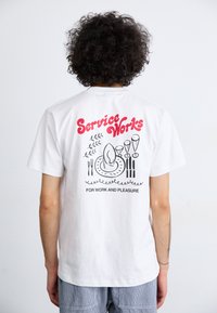 Camiseta de algodón blanca con un diseño gráfico negro que presenta un plato, utensilios y el texto "Service Works" en rojo en la parte de atrás.