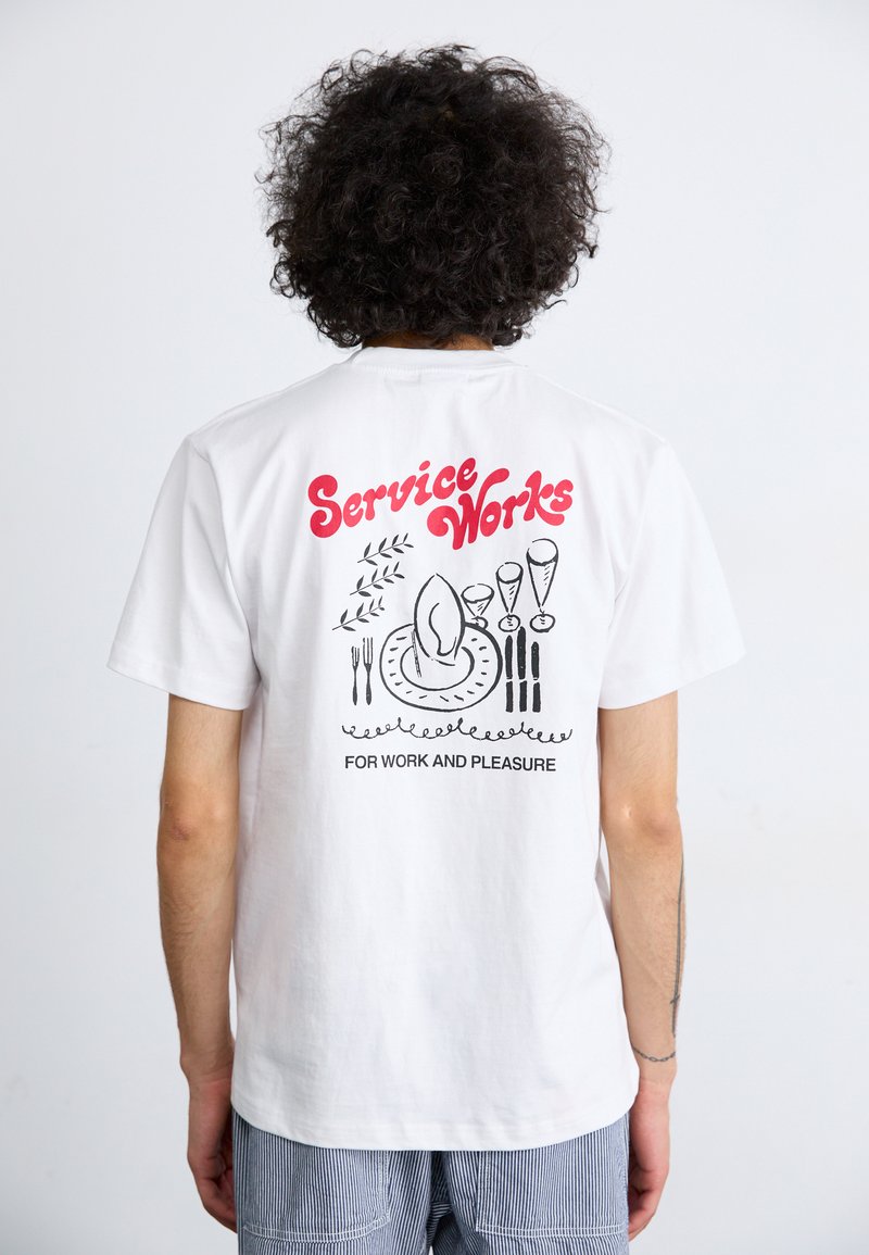 Camiseta de algodón blanca con un diseño gráfico negro que presenta un plato, utensilios y el texto "Service Works" en rojo en la parte de atrás.