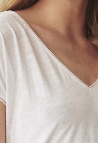Jasnoróżowa koszulka z dekoltem w kształcie litery V i krótkimi rękawami, wykonana z miękkiego, teksturowanego materiału. Charakteryzuje się delikatnym przeszyciem wzdłuż dekoltu i brzegów.