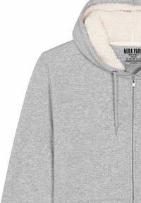 Felpa grigia con zip e una morbida fodera in pile bianco. Presenta lacci e una cerniera liscia. L'etichetta include il nome del marchio e le istruzioni per la cura.