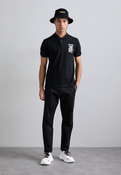 Black polo shirt with a bear graphic, paired with black pants and white sneakers. The outfit includes a black bucket hat with gold text.