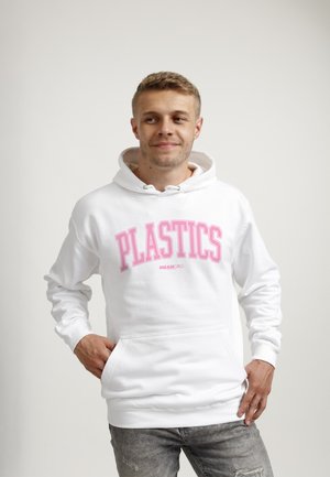 Junger Mann trägt weißen Hoodie mit pinkem Schriftzug „PLASTICS MEAN GIRLS“, steht mit Händen in den Taschen, schaut zur Seite und lächelt leicht.