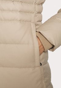 Beige puffjas met een gepatineerd ontwerp en een zijzak. Gebreide manchet zichtbaar eronder. Zachte, gladde textuur. Klein zwart logo detail.