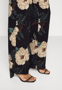 Kwiatowe spodnie szerokie w nogawkach z dużymi beżowymi piwoniami na czarnym tle, zestawione z czarnymi płaskimi sandałami z cienkimi paskami.