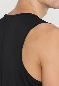 Detailní záběr na rameno a horní část zad muže, který má na sobě černé sportovní tílko bez rukávů z texturované látky.