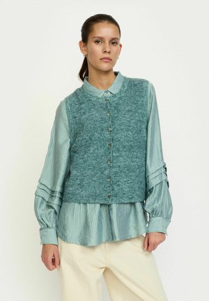 Vestă tricotată teal cu decolteu rotund și nasturi frontali, asortată cu o cămașă albastru deschis texturată cu mâneci lungi; pantaloni de culoare crem.