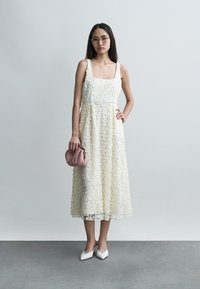 Γυναίκα με μεσομήκους φόρεμα από κρεμ δαντέλα με λουλούδια, λευκά μυτερά τακούνια, κρατώντας μια μικρή ροζ τσάντα, στέκεται μπροστά σε απλό γκρι φόντο.