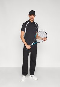 Μαύρο πόλο πουκάμισο με λευκές λεπτομέρειες και λογότυπο, μαύροι υφασμάτινοι παντελόνια με υφή και λευκά αθλητικά παπούτσια. Κρατάει ρακέτα τένις. Μαύρο καπέλο.