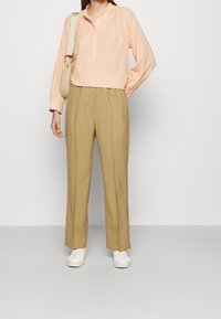 Person trägt eine pfirsichfarbene Langarmbluse mit Knöpfen, beigem High-Waist-Hose, weißen Turnschuhen und hält eine beige Umhängetasche, während sie vor einer weißen Wand steht.
