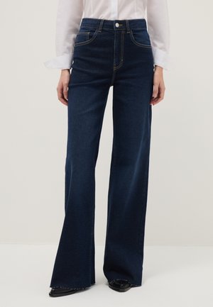 Femme portant un jean denim bleu foncé à jambes larges, associé à une chemise blanche à manches longues et des chaussures noires à bout pointu.