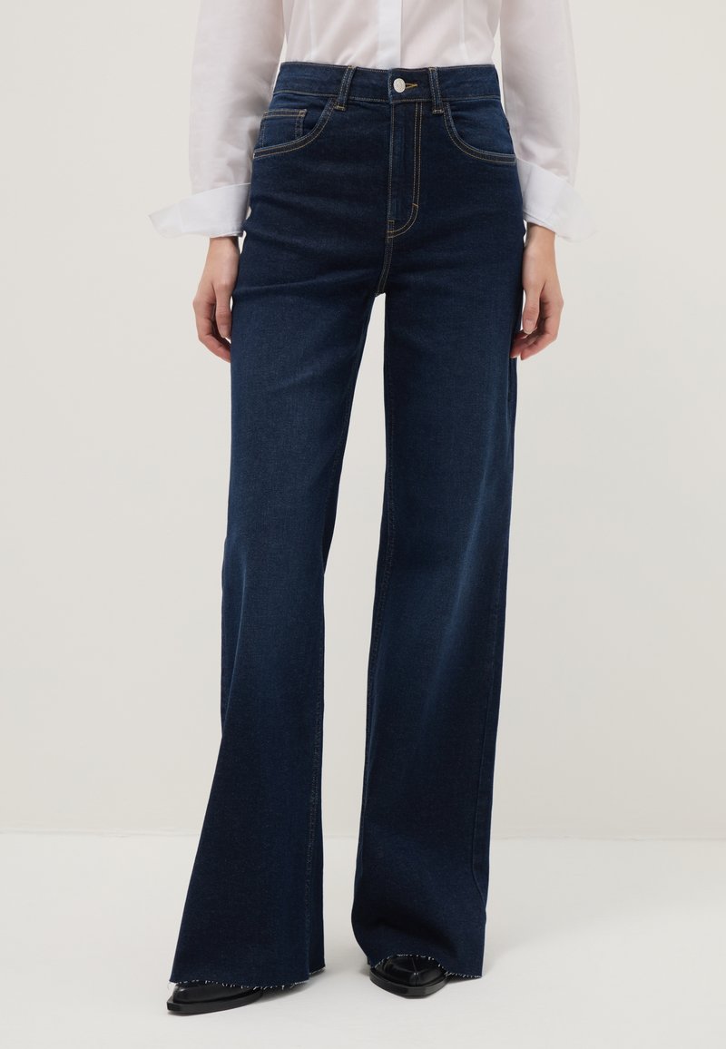 Femme portant un jean denim bleu foncé à jambes larges, associé à une chemise blanche à manches longues et des chaussures noires à bout pointu.