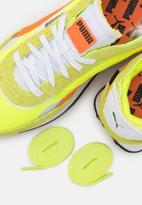 Puma-Sneaker mit einem leuchtend gelben und grünen Obermaterial aus Wildleder, orangefarbenen Akzenten, weißen Schnürsenkeln und zwei weiteren neon-gelben Schnürsenkeln.