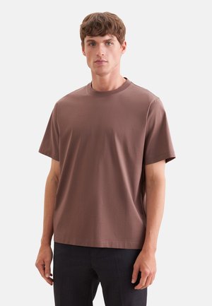 Jonge man met kort bruin haar, die een loszittend bruin T-shirt met korte mouwen en een donkere broek draagt, staand tegen een effen achtergrond.