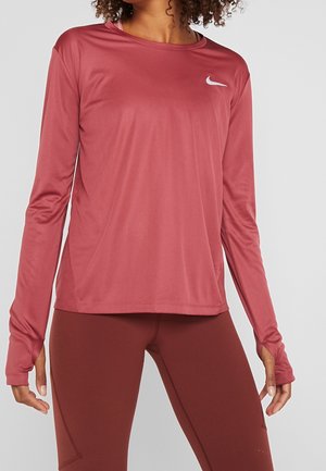 Femme portant un haut de sport à manches longues rose et des leggings assortis, debout devant un fond gris clair.