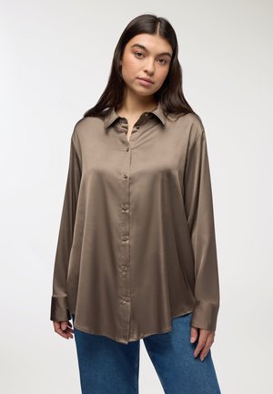 Camicia in seta khaki con colletto, maniche lunghe e chiusura frontale con bottoni. Tessuto liscio e vestibilità comoda. Pantaloni in denim blu visibili.