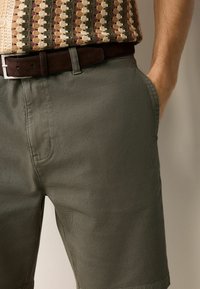 Olivenfarvede shorts med en tekstureret overflade, der har sidelommer og et brunt ruskindsbælte. Stoffet er vævet med et holdbart design.