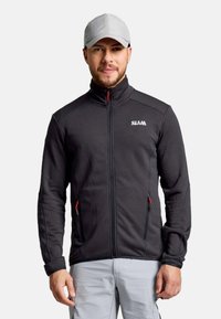 Mann in einer schwarzen Zip-Jacke mit zwei Reißverschlusstaschen und einer hellgrauen Mütze, stehend vor einem schlichten hellen Hintergrund.