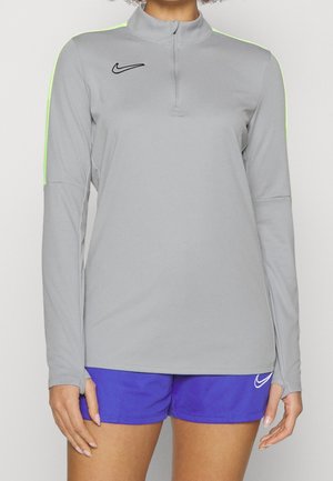 Persoon draagt een lichtgrijs atletisch shirt met lange mouwen met neon groene strepen en blauwe sportshorts met logo's.