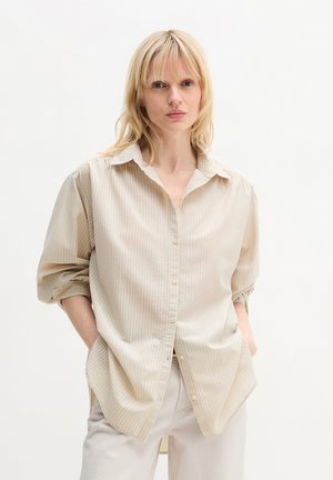 Blonde vrouw met steil haar, gekleed in een beige gestreept overhemd met knopen en lichtgekleurde broek, staand met handen in de zakken tegen een witte achtergrond.