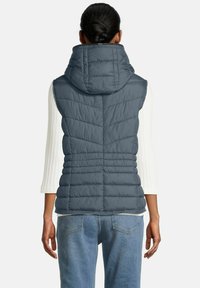 Vrouw met donker haar in een bun, gekleed in een blauwe gewatteerde hoodie-vest over een witte lange mouwen top en blauwe jeans, weergegeven van achter.