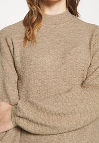 Beżowy, strukturalny sweter z dzianiny o wysokim dekolcie i długich rękawach. Posiada subtelny wzór oraz ściągacze, zapewniając wygodne dopasowanie.