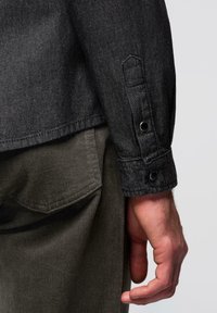 Chemise en jean noire avec un poignet comportant deux boutons-pression en métal. Un pantalon en velours côtelé vert olive est partiellement visible en arrière-plan.
