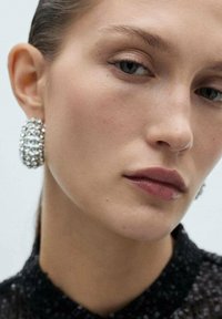 Sølv, runde øreringe med et tekstureret design besat med krystaller. Modellen har glat hud og neutral makeup. Baggrunden er ensfarvet.