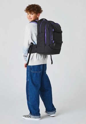 Kind trägt einen großen schwarzen und lila Rucksack, einen lässigen Pullover, weite Jeans und karierten Slip-On-Schuhen, steht und schaut über die Schulter.