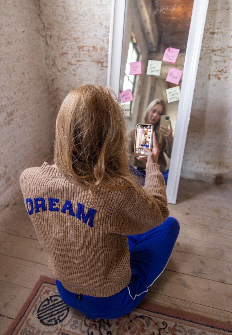 Beige gebreide trui met "DREAM" in het blauw op de achterkant, gecombineerd met blauwe joggingbroeken. Een spiegel weerspiegelt een persoon die een smartphone vasthoudt.
