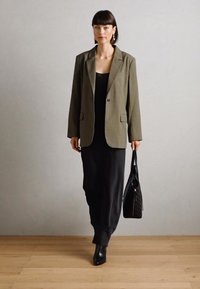 Blazer surdimensionné vert olive sur une robe noire, avec un sac à main noir texturé. Le mannequin porte des bottines noires ; design minimaliste et couleurs à fort contraste.