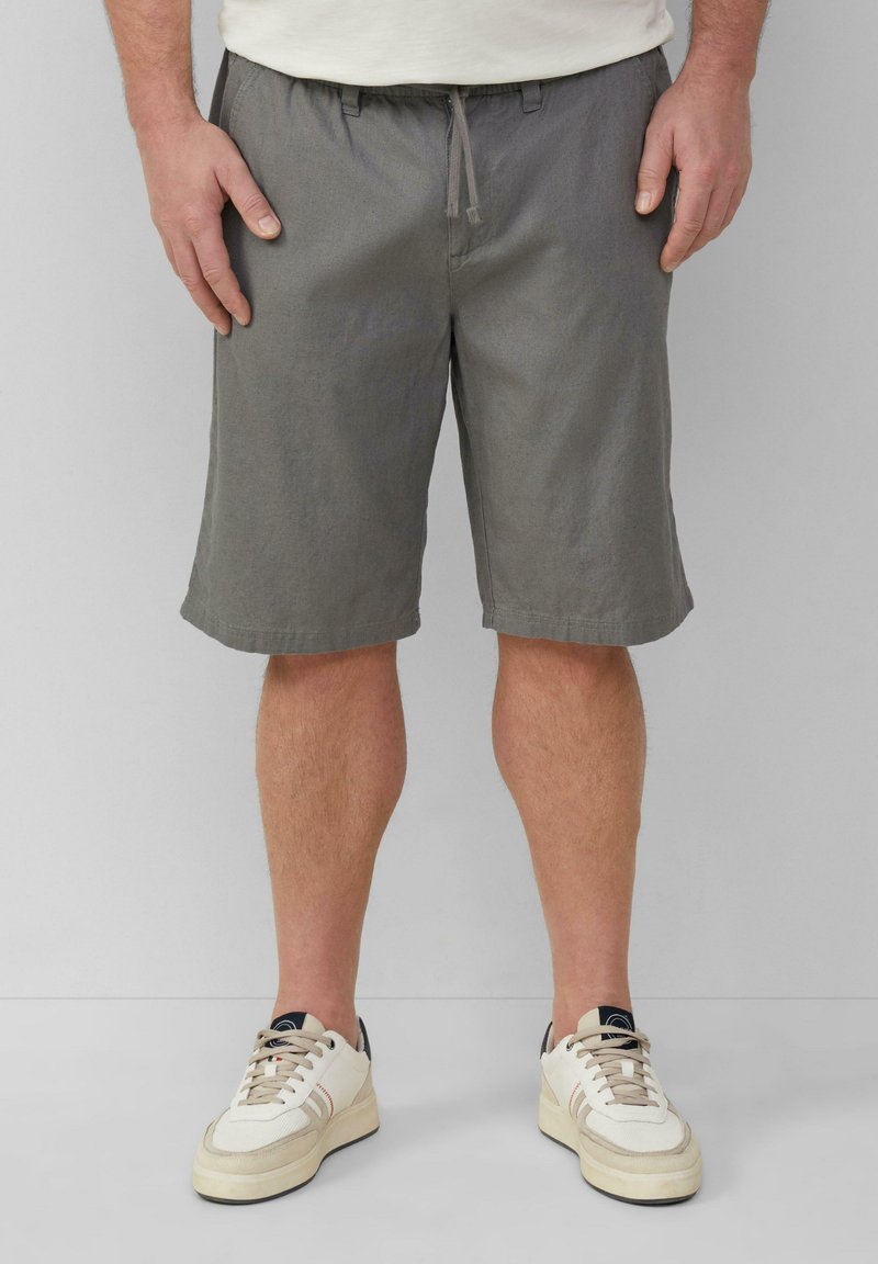 Homme portant un short gris jusqu'aux genoux et des baskets blanches, se tenant devant un fond gris clair uni.