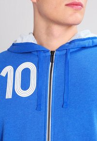 Sweat à capuche bleu zippé avec un numéro "10" blanc imprimé sur le devant. Comprend une capuche standard avec cordons et un tissu texturé.
