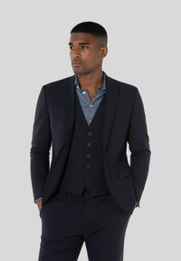 Mężczyzna stojący z rękami w kieszeniach, ubrany w ciemnogranatowy garnitur, kamizelkę i niebieską koszulę zapinaną na guziki na jednolitym szarym tle.