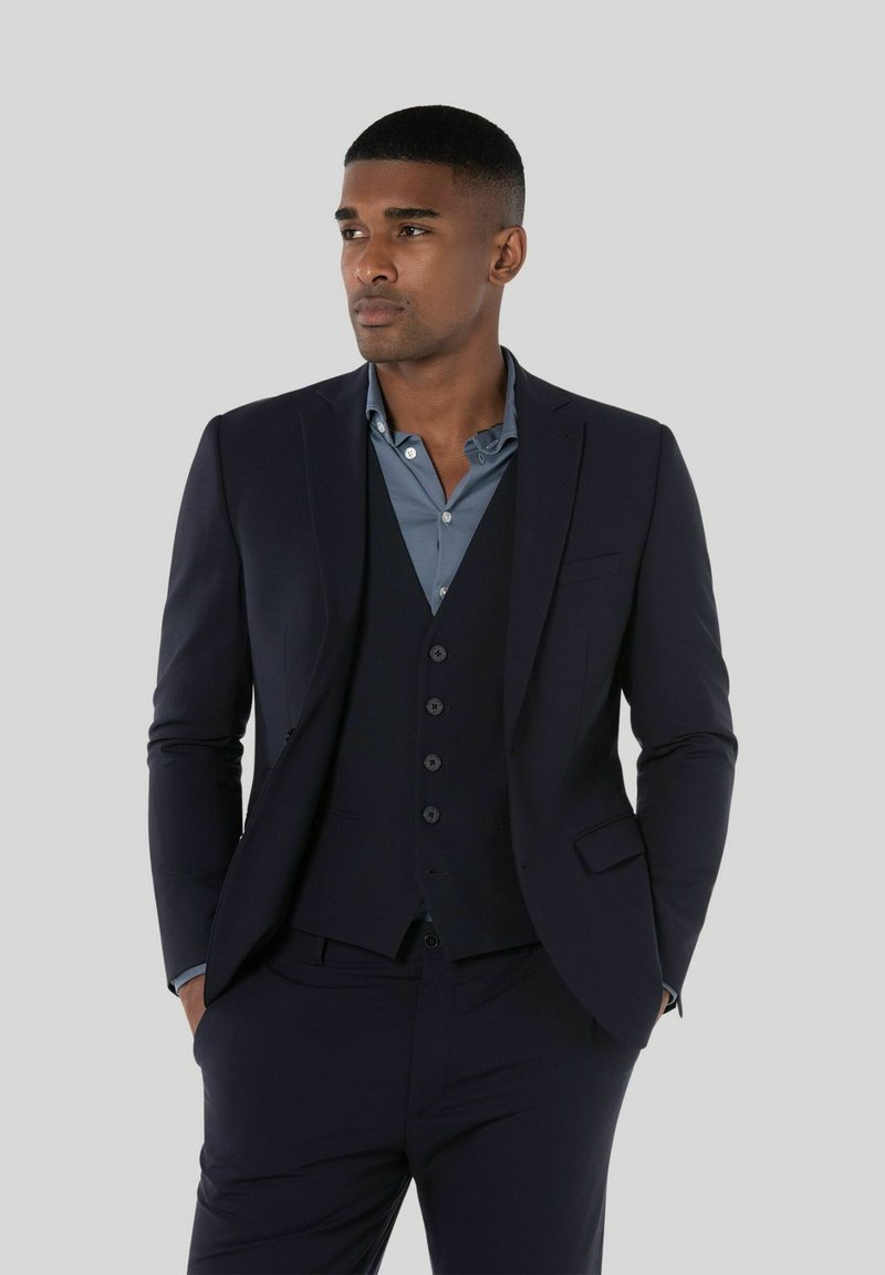 Mężczyzna stojący z rękami w kieszeniach, ubrany w ciemnogranatowy garnitur, kamizelkę i niebieską koszulę zapinaną na guziki na jednolitym szarym tle.