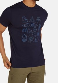 Man med marinblå skjorta prydd med ljusblå campingikoner som inkluderar stövlar, kanna, kajak, träd, ryggsäck, sol, tält, kompass, lägereld och rep.