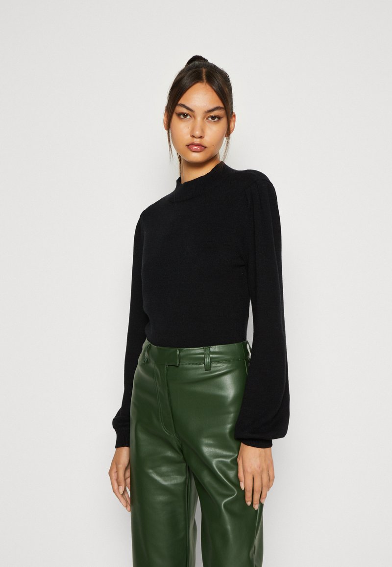 Black knit sweater with a high neckline and puffed long sleeves, paired with high-waisted green faux leather trousers.