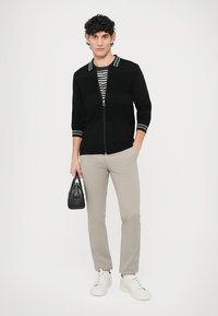 Černý svetr na zip s pruhovaným límcem, kombinovaný s pruhovaným tričkem, světle béžové kalhoty a bílé tenisky. Drží malou černou tašku.