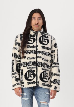 Fleece jacket in cream with black lettering. Features a front zipper, stand collar, and relaxed fit. Text includes "Gues" and graphic elements.