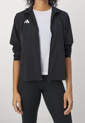 Personne portant une veste noire zippée sur un t-shirt sportif blanc et un legging noir, debout les bras détendus le long du corps.