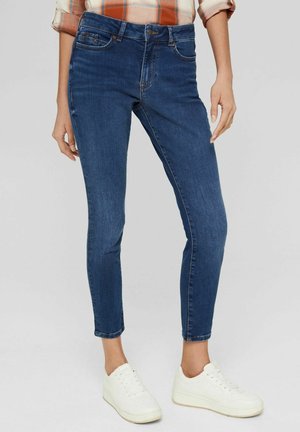 Femme portant un jean skinny bleu foncé, une chemise à carreaux orange avec les manches retroussées, et des baskets blanches, debout devant un fond neutre.