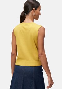 T-shirt en tricot jaune à manches courtes avec ourlet côtelé. Porté avec une jupe en denim, présenté de dos, mettant en valeur l'encolure et les ouvertures des bras.