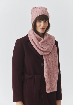 Berretto e sciarpa rosa chiaro lavorati a maglia con un motivo a foglia testurizzato, indossati con un cappotto bordeaux scuro. Il berretto aderisce bene, la sciarpa cade morbida.