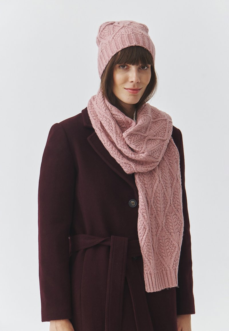 Világos rózsaszín kötött sapka és sál, texturált levélmintával, sötét bordó kabáttal viselve. A sapka szorosan illeszkedik, a sál lazán omlik.