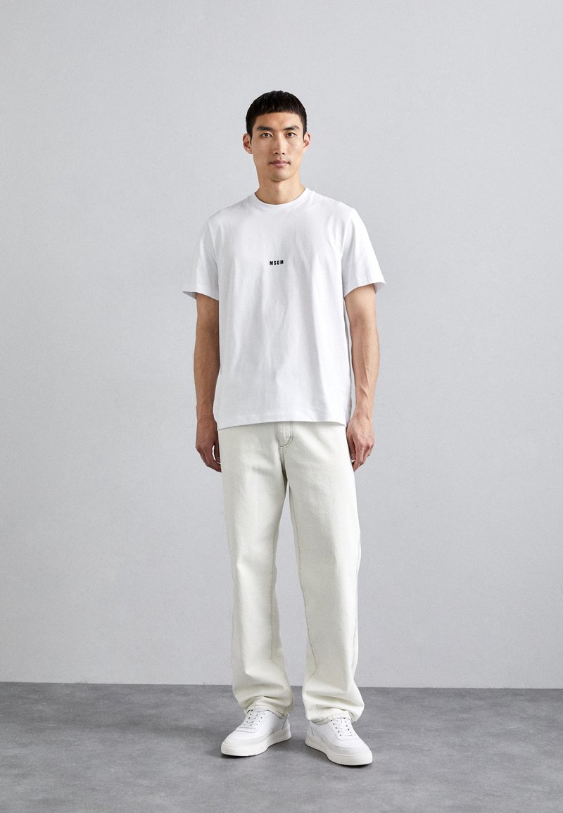 White crew-neck t-shirt with a black logo, paired with off-white straight-leg jeans and white sneakers, standing against a neutral backdrop.
