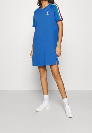 Femme portant une robe courte bleue Adidas avec le logo de l'Italie et des bandes vertes, blanches et rouges sur les épaules, assortie à des baskets et des chaussettes blanches.